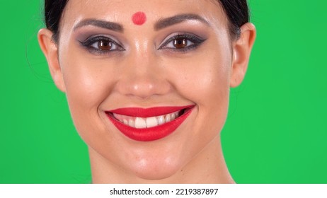 Portrait Of Beautiful Indian Woman With Red Dot On Forehead Looking At Camera And Smiling Revealing Teeth. Close Up Of A Brunettes Face With Bright Eye Makeup And Red Lips On Green Screen At Studio.