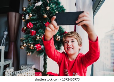 Portrait Of Beautiful Kid Boy At Home By The Christmas Tree And Using Mobile Phone To Take A Selfie