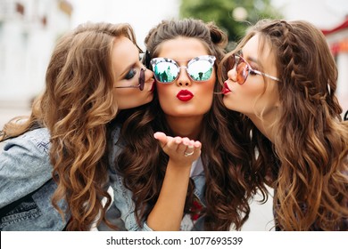 Portrait of beautiful ladies with hairstyles and make up wearing stylish sunglasses and showing peace signs while kissing friend in mirrored sunglasses with red lisp showing heart shaped gesture at ca