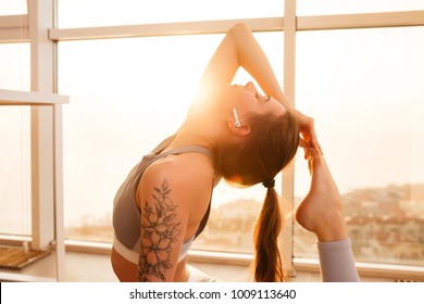 Portrait of beautiful lady practicing yoga poses on yoga mat while listening music in earphones at home with big beautiful windows on background