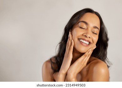 Portrait of beautiful latin woman touching her clean and healthy face against grey background. Smiling hispanic woman with natural makeup feeling healthy skin with eyes closed. Beauty care pampering.