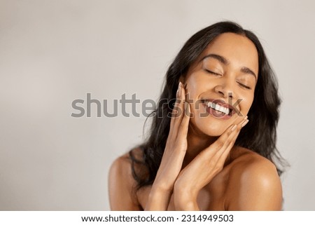 Portrait of beautiful latin woman touching her clean and healthy face against grey background. Smiling hispanic woman with natural makeup feeling healthy skin with eyes closed. Beauty care pampering.