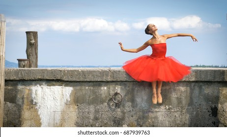 Portrait of a beautiful light ballerina, in a lush red dress, sits on a stone pier, tender. Concept: ballet dancer, slim, diet, light women, flexibility, beautiful figure, healthy lifestyle, sky. - Powered by Shutterstock