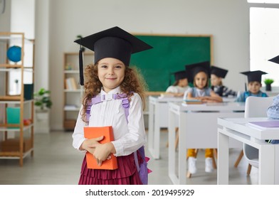 Portrait of a beautiful little curly elementary school graduate girl wearing an academic hat. Child with a book in his hands stands in the classroom and looks at the camera. Education concept. - Powered by Shutterstock