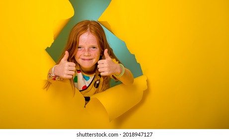 Portrait of beautiful little ginger kid girl giving you thumbs up over yellow background. breaking through yellow paper wall. Happy smiling kid go back to school