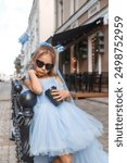 portrait of beautiful little girl in blue poofy dress and sunglasses with coffee standing in city. Fashion kid