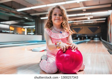 Portrait of a beautiful little girl with freckles smiling and holding a pink bowling ball, sitting on a bowling track.