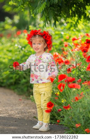 Portrait of a beautiful little girl having fun in field of red poppy flowers in spring. Lovely smiling child girl with blond hair and a wreath of real flowers on her head.