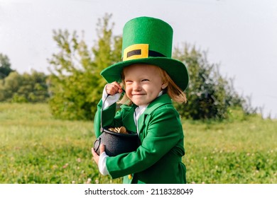 Portrait of a beautiful little girl in a leprechaun costume who holds a pot of gold coins in her hands. Leprinon embraces his gold in a large green meadow with clover. The child in the hat smiles