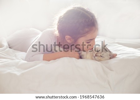 Portrait of a beautiful little girl, lies on a clean, white bed with a white British cat.