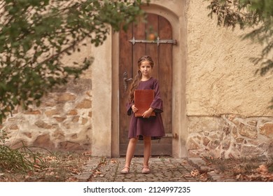 Portrait Of A Beautiful Little Girl With An Old Encyclopedia In Her Hands In The Courtyard Of The House.