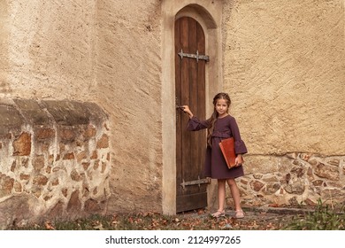 Portrait Of A Beautiful Little Girl With An Old Encyclopedia In Her Hands In The Courtyard Of The House.