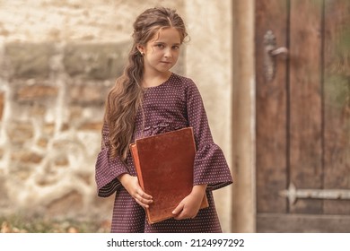 Portrait Of A Beautiful Little Girl With An Old Encyclopedia In Her Hands In The Courtyard Of The House.