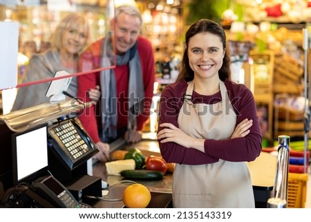Portrait of beautiful long-haired young woman cashier smiling at camera at workplace. Friendly worker assisting cheerful senior couple at cashdesk at newest supermarket. Good service concept