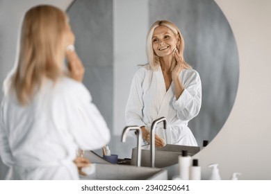 Portrait Of Beautiful Mature Lady Cleansing Skin With Cotton Pad Near Mirror In Bathroom, Attractive Senior Woman Smiling To Her Reflection, Enjoying Self Care Routine At Home, Selective Focus