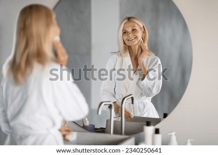 Portrait Of Beautiful Mature Lady Cleansing Skin With Cotton Pad Near Mirror In Bathroom, Attractive Senior Woman Smiling To Her Reflection, Enjoying Self Care Routine At Home, Selective Focus