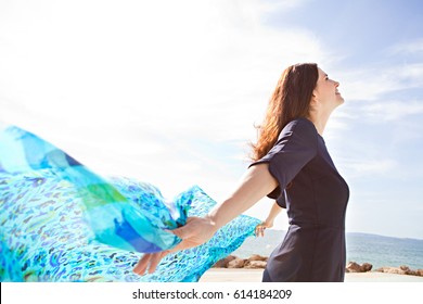 Portrait Of A Beautiful Mature Woman Breathing Fresh Air By The Sea, Holding A Floating Fabric Scarf In The Breeze On Sunny Day, Outdoors. Well Being And Healthy Lifestyle, Dreamy Positive Exterior.