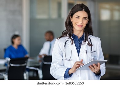 Portrait of beautiful mature woman doctor holding digital tablet and looking at camera. Confident female doctor using digital tablet with colleague talking in background at hospital. Latin nurse.