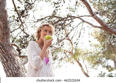 Portrait Of Beautiful Mature Woman In Nature With Trees Biting A Green Apple On A Sunny Day, Outdoors. Healthy Senior Woman Eating Fruit, Wellness And Well Being Lifestyle, Smiling In Sunny Exterior.