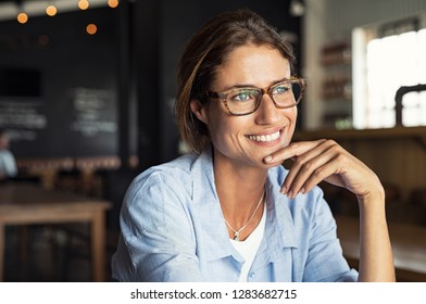 Portrait of beautiful mature woman sitting in cafeteria looking away. Cheerful mature woman wearing eyeglasses thinking with finger on chin. Happy woman relaxing at cafe and smiling.