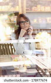 Portrait Of Beautiful Mature Woman Sitting In Coffee Shop And Using Her Mobile. Shot Through Window.