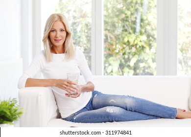 Portrait Of Beautiful Mature Woman Smiling While Sitting At Sofa At Home And Drinking A Glass Of Water. 