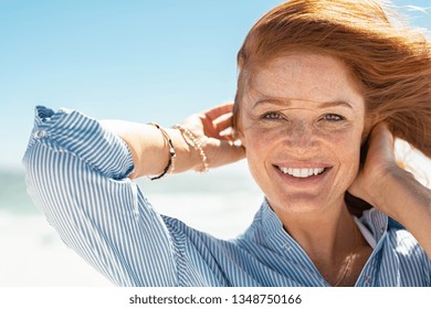 Portrait Of Beautiful Mature Woman With Wind Fluttering Hair. Closeup Face Of Healthy Young Woman With Freckles Looking At Camera. Lady With Red Hair Standing At Seaside Enjoying Breeze At Beach.