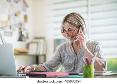 the portrait of a beautiful  middle age woman, sitting at her desk in her home. she is talking at cell phone and is using her laptop. there is also a tablet on the desk