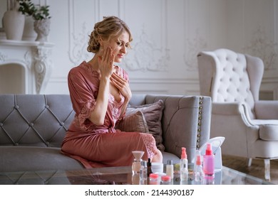 Portrait beautiful middle aged woman looking in mirror sitting on cozy sofa at home, attractive female touching neck skin posing for phot, natural beauty and skin care sitting on sofa in pink bathrobe