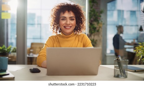 Portrait of a Beautiful Middle Eastern Manager Sitting at a Desk in Creative Office. Young Stylish Female with Curly Hair Looking at Camera with Big Smile. Colleagues Working in the Background. - Shutterstock ID 2231710223
