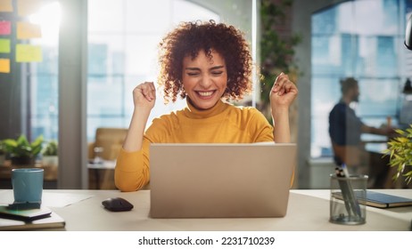 Portrait of a Beautiful Middle Eastern Manager Sitting at a Desk in Creative Office. Young Stylish Female with Curly Hair Celebrating her Achievement with Big Smile - Powered by Shutterstock