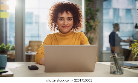 Portrait of a Beautiful Middle Eastern Manager Sitting at a Desk in Creative Office. Young Stylish Female with Curly Hair Looking at Camera with Big Smile. Colleagues Working in the Background. - Powered by Shutterstock