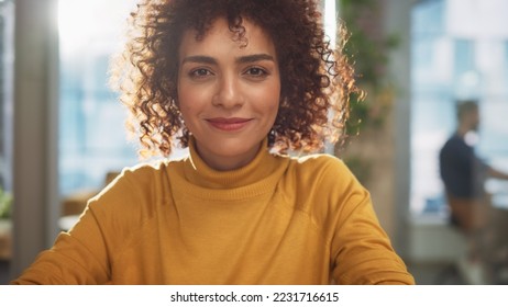 Portrait of a Beautiful Middle Eastern Manager Sitting at a Desk in Creative Office. Young Stylish Female with Curly Hair Looking at Camera with Big Smile. Colleagues Working in the Background. - Powered by Shutterstock