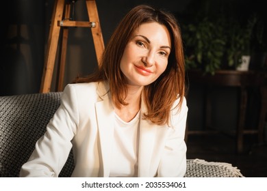 Portrait Of A Beautiful Middle-aged Woman With Straight Shoulder-length Hair. A Woman Sits In A Chair In A Cabinet With Beautiful Natural Light. Easy Smile On A Woman's Face Evokes Sympathy And Trust