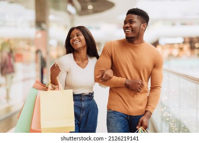 Portrait of beautiful millennial African American guy and lady shopping on weekend, walking in the city mall with colorful bags, looking at boutique window and laughing spending time together