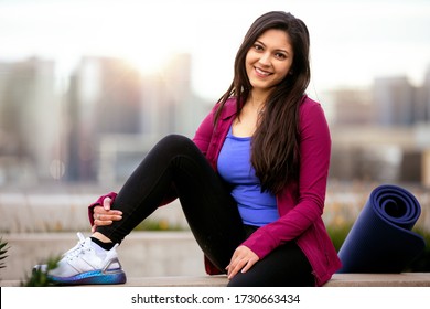 Portrait of a beautiful mixed ethnicity brunette woman in sportswear, sitting relaxed after a morning yoga workout with city skyline in the background
