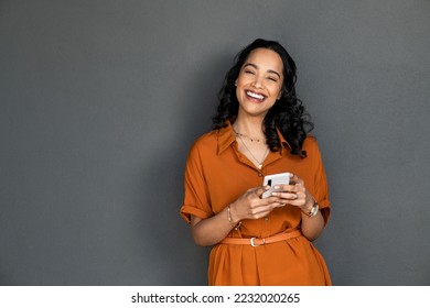 Portrait of beautiful mixed race woman standing against grey wall using smartphone. Cheerful latin young woman holding mobile phone while looking at camera. Happy carefree woman messaging. - Shutterstock ID 2232020265