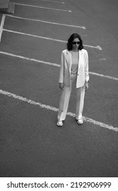 Portrait Of Beautiful Modern Woman In Parking Area. Fashionable. White Suit. Crop Top. Black Hair. Slim And Tall. Daily Life. Work And Leisure. City Background. Black And White Photo