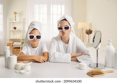 Portrait Of Beautiful Mother And Daughter Wearing Cool Trendy Shades And White Bath Towels Duck Facing Sitting At Table With Mirror And Beauty Products On Spa Day Or During Morning Skincare Routine