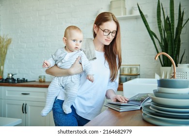 Portrait Of Beautiful Mother With Her Cute Little Baby Looking Through A Cook Book In A Cozy Kitchen At Home. Kitchen Design, Furniture For Kitchen. Happy Modern Family. 