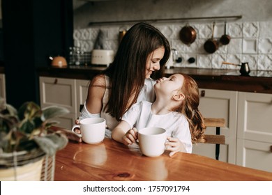 Portrait Of A Beautiful Mother And Her Little Girl Sitting At Home And Sharing A Happy Moment Together In A Dark Kitchen. A Young Mother And A Small Red-haired Daughter Holds A Cup Of Tea In Her Hand.