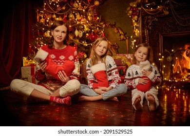 Portrait Of Beautiful Mother And Her Sweet Daughters Sitting In A Row By A Xmas Tree And Fireplace With Stockings Filled With Gifts. Cozy Beautiful Decorated Room Background. Happy Christmas Family. 