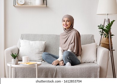 Portrait of beautiful muslim girl in hijab sitting on sofa at home and looking at camera, resting in stylish living room interior