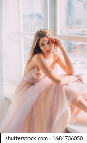 Portrait Beautiful Mysterious Girl With Long Blond Hair In Luxurious Airy Elegant Pastel Beige Dress Natural Cosmetics. Princess Sits On Windowsill, Hand Touches Head Backdrop White Window. Prom Party