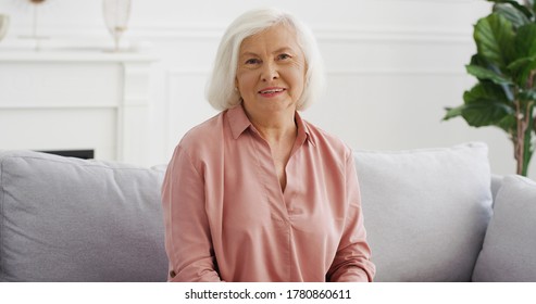 Portrait of beautiful old Caucasian woman with gray hair sitting on couch in living room and talking to camera. Female vlogger telling something and explaining. Video blogging Vlog from home Videochat