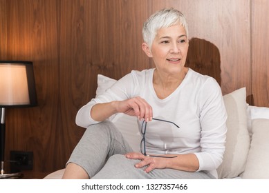 Portrait Of Beautiful Pensive Senior Woman With Glasses Sitting On A Comfy Bed And Getting Ready For Sleep In Her Bright Modern Bedroom, Night Routine And Bedtime Concept