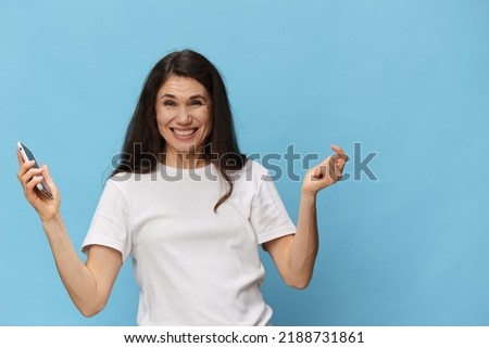 portrait of a beautiful, pleasant woman with long hair, in a light T-shirt, holding a fashionable smartphone in her hands and joyfully raising her hands up. Horizontal photo on a light blue
