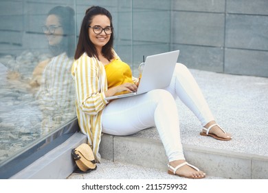 Portrait Of Beautiful Plus Size Woman 30-35 Years Old In Fashion Clothes And Eyeglasses Working Remotely On A Laptop. Female Freelancer Sitting On The Floor, Typing On A Computer In The Park Outdoors