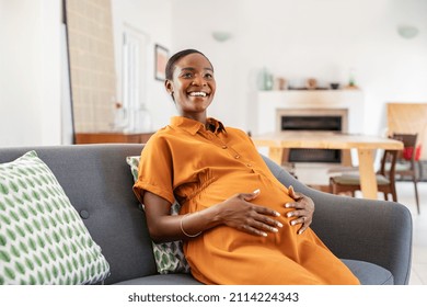 Portrait of beautiful pregnant woman relaxing at home while touching belly. Smiling middle aged african american woman sitting on couch. Happy black pregnant woman resting and touching her tummy.