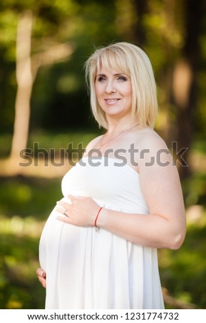 Portrait of beautiful pregnant woman in sheer long white maternity dress looking dreamy in the middle of forest in sunny summer day.